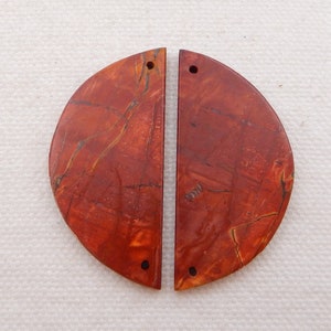 Puede incluir: Dos cuentas de piedra jaspe rojo en forma de semic&iacute;rculo con agujeros perforados. Las cuentas tienen un patr&oacute;n de m&aacute;rmol rojo, naranja y marr&oacute;n.