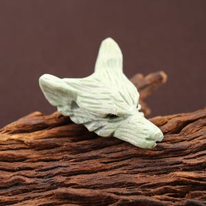 May include: A pale green stone carving shaped like a fox head. The sculpture features detailed etched lines representing fur texture, pointed ears, and small dark eyes. The fox head is oriented to the right, showcasing a sharp snout and facial features.