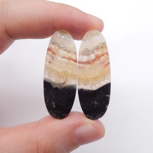 May include: Two oval-shaped gemstone pendants. The pendants have a layered appearance with bands of black, beige, orange, and white. Each pendant has a small hole at the top for stringing. The gemstones are held by a hand.