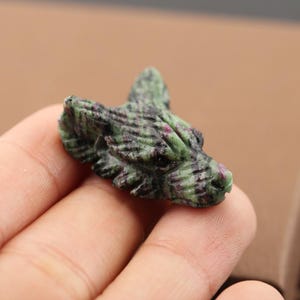 May include: A small, hand-carved stone figurine in the shape of a wolf head. The sculpture is crafted from a mottled green and dark red mineral, featuring detailed textures that mimic fur and pointed ears, with a smooth, polished finish throughout.