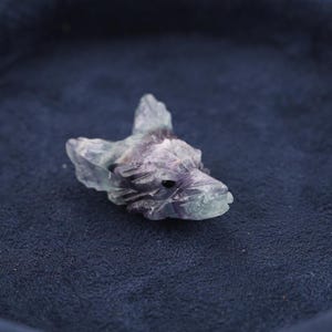 May include: A carved fluorite gemstone shaped like a wolf head, featuring translucent layers of purple, green, and clear mineral tones. The sculpture displays detailed facial features, including pointed ears and a snout, with a polished finish.