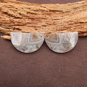 May include: Two semi-circular, polished agate stones with a natural, layered pattern in shades of white, gray, and brown. Each stone has a small drilled hole. The stones are displayed on a brown surface with a piece of textured wood.