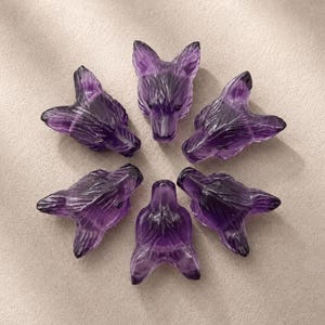 May include: Six individual wolf head carvings made from deep purple amethyst crystal. Each piece features detailed etched fur textures and pointed ears, arranged in a circular pattern with the faces pointing outward from the centre point.