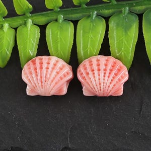 Op de afbeelding: Twee roze schelpvormige kralen met een gedetailleerde textuur. De kralen zijn gemaakt van een glad, glanzend materiaal.