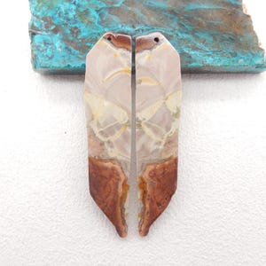 May include: Two polished gemstone pendants with a light pink and cream color, featuring brown and yellow veining. The bottom portion of each pendant is a rich brown color. Each pendant has a small hole at the top.