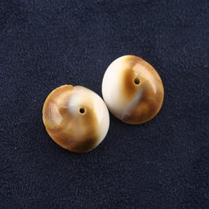 May include: Two polished, dome-shaped shell beads featuring a smooth, glossy surface with a gradient of creamy white transitioning into a rich, warm brown. Each bead has a small, centered circular hole, showcasing a natural, organic aesthetic.