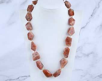 May include: A necklace featuring large, irregular, faceted sunstone beads in shades of reddish-brown with natural sparkling inclusions. The stones are strung together in a single strand, showcasing a raw, geometric shape and a polished finish.