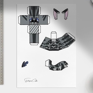 May include: A papercraft cat template with various cut-out pieces for assembly. The cat design features gray and black stripes, with blue eyes and pink inner ears. A ruler is on the left side of the image.