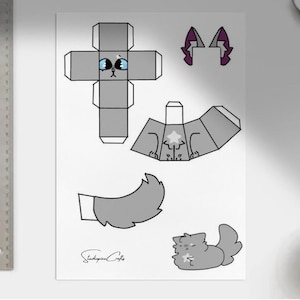 May include: A sheet of paper with a gray cat-shaped papercraft design. The design includes a box-shaped head with blue eyes, body, tail, and ears. The design is accompanied by a ruler and a cup of coffee.