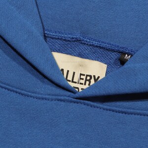 May include: Close-up of a blue hoodie's neckline. The interior tag reads "GALLERY" in black letters. The hoodie is a solid blue color, with a soft, textured appearance. The tag also has the letter "M" on it.
