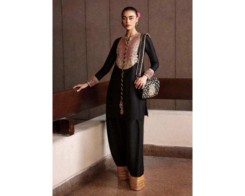K&ouml;nnte beinhalten: Schwarzes Ensemble mit gold- und rosafarbenen Stickereien auf dem Oberteil und den Manschetten. Das Outfit umfasst eine passende schwarze Hose und eine dekorative Umh&auml;ngetasche mit goldenen Details. Die Hose hat einen gold-orangenen Saum.
