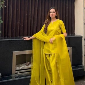 K&ouml;nnte beinhalten: Ein leuchtend gelbes zweiteiliges Outfit. Das Oberteil ist eine kurz&auml;rmelige Tunika mit rundem Ausschnitt. Die passende Hose ist weit geschnitten. Ein durchsichtiger Schal drapiert &uuml;ber den Schultern. Das Outfit ist aus glattem, gl&auml;nzendem Stoff.