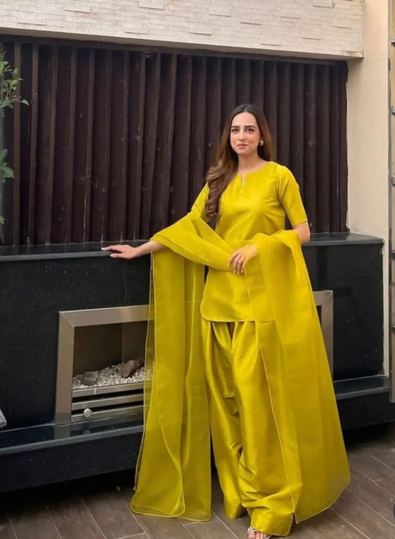 K&ouml;nnte beinhalten: Ein leuchtend gelbes zweiteiliges Outfit. Das Oberteil ist eine kurz&auml;rmelige Tunika mit rundem Ausschnitt. Die passende Hose ist weit geschnitten. Ein durchsichtiger Schal drapiert &uuml;ber den Schultern. Das Outfit ist aus glattem, gl&auml;nzendem Stoff.