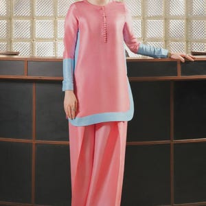 May include: A pink two-piece outfit with light blue accents. The long-sleeved top has a button-up front and light blue cuffs. The wide-leg pants have light blue trim at the hem. The outfit is worn by a person with glasses.