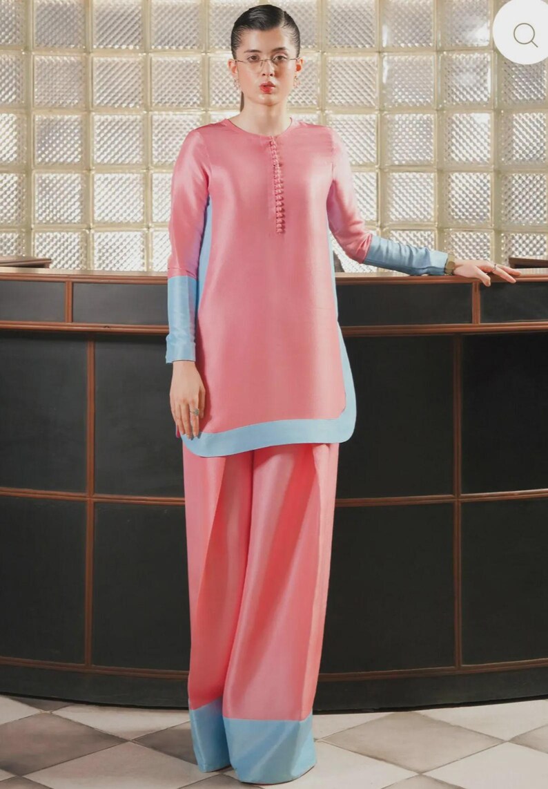May include: A pink two-piece outfit with light blue accents. The long-sleeved top has a button-up front and light blue cuffs. The wide-leg pants have light blue trim at the hem. The outfit is worn by a person with glasses.