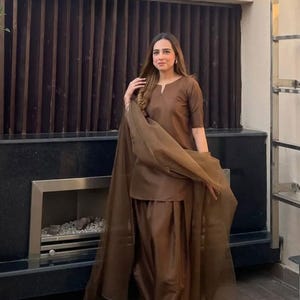 K&ouml;nnte beinhalten: Ein braunes Salwar-Kameez-Outfit. Das Ensemble umfasst ein Tunika-Oberteil mit V-Ausschnitt, eine passende Hose und einen transparenten Schal. Das Outfit hat eine einfarbige, warme braune Farbe. Der Hintergrund zeigt eine dunkelbraune Lamellenwand.