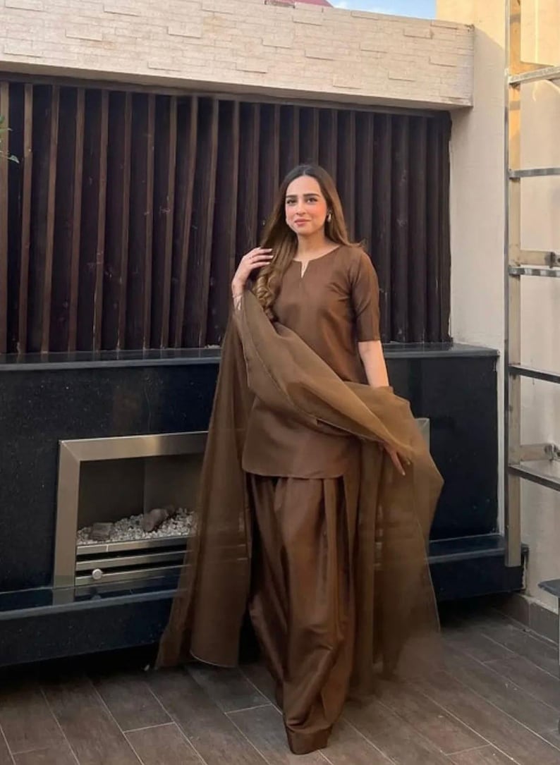 K&ouml;nnte beinhalten: Ein braunes Salwar-Kameez-Outfit. Das Ensemble umfasst ein Tunika-Oberteil mit V-Ausschnitt, eine passende Hose und einen transparenten Schal. Das Outfit hat eine einfarbige, warme braune Farbe. Der Hintergrund zeigt eine dunkelbraune Lamellenwand.