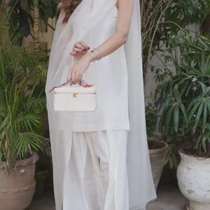 K&ouml;nnte beinhalten: Ein wei&szlig;es Ensemble aus einem Oberteil mit hohem Kragen, weiten Hosen und einem transparenten Cape. Eine kleine, rechteckige wei&szlig;e Handtasche mit goldenem Verschluss wird gehalten. Die Szene ist ein Garten mit Topfpflanzen.