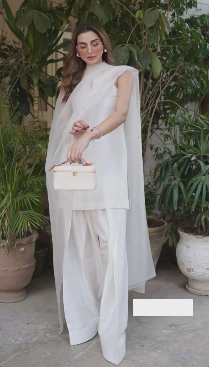 K&ouml;nnte beinhalten: Ein wei&szlig;es Ensemble aus einem Oberteil mit hohem Kragen, weiten Hosen und einem transparenten Cape. Eine kleine, rechteckige wei&szlig;e Handtasche mit goldenem Verschluss wird gehalten. Die Szene ist ein Garten mit Topfpflanzen.