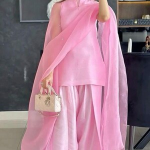 K&ouml;nnte beinhalten: Hellrosa Outfit bestehend aus einer Tunika, einer Hose mit weitem Bein und einem transparenten Schal. Das Outfit wird mit einer kleinen, cremefarbenen Handtasche und silbernen Sandalen kombiniert. Das Ensemble ist elegant und f&uuml;r besondere Anl&auml;sse geeignet.