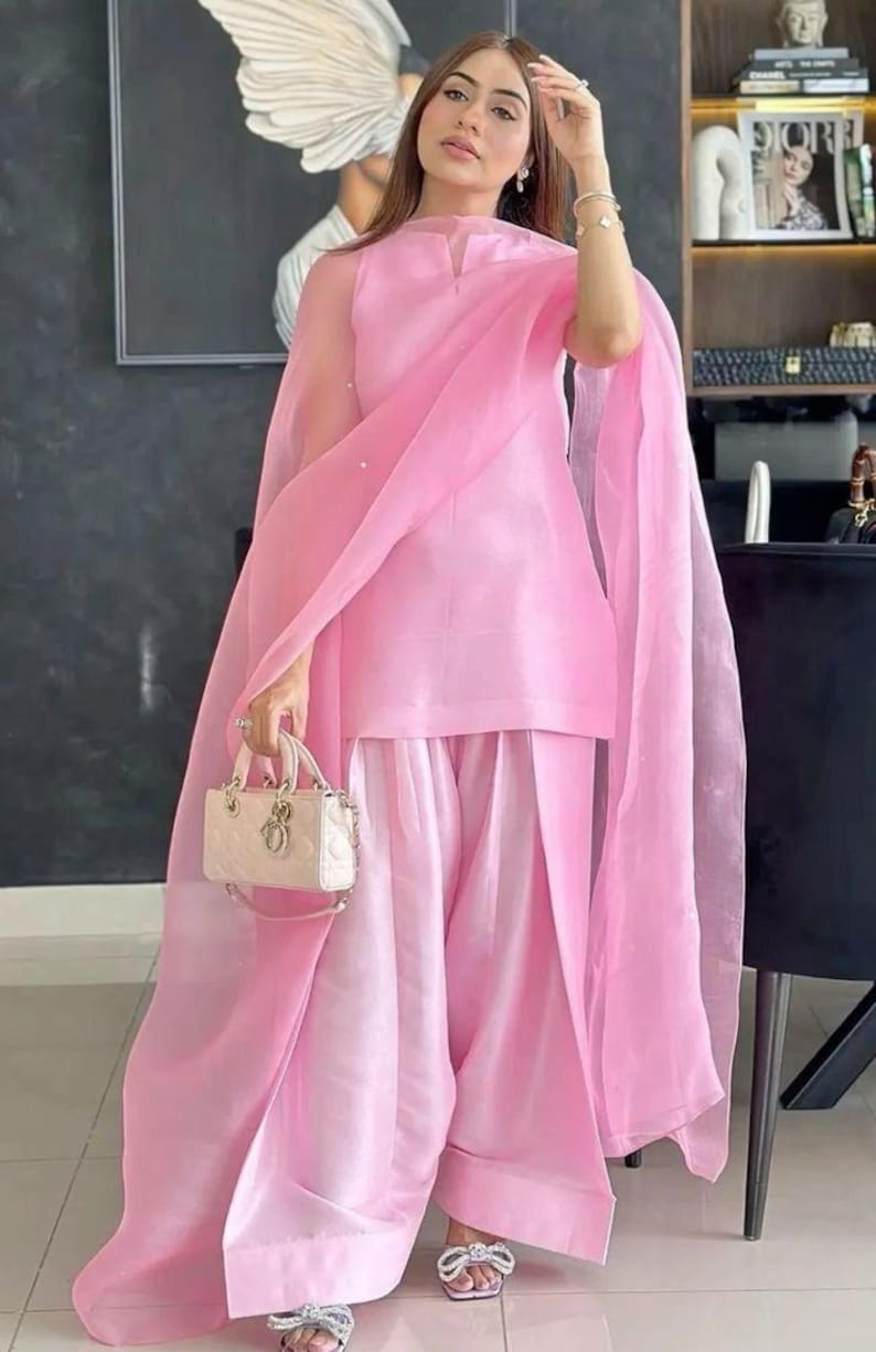 K&ouml;nnte beinhalten: Hellrosa Outfit bestehend aus einer Tunika, einer Hose mit weitem Bein und einem transparenten Schal. Das Outfit wird mit einer kleinen, cremefarbenen Handtasche und silbernen Sandalen kombiniert. Das Ensemble ist elegant und f&uuml;r besondere Anl&auml;sse geeignet.