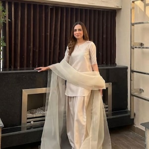 K&ouml;nnte beinhalten: Beigefarbenes zweiteiliges Outfit mit passendem, transparentem Schal. Das Outfit umfasst ein kurz&auml;rmeliges Oberteil und eine locker sitzende Hose. Der Schal ist &uuml;ber den Schultern drapiert. Das Outfit ist aus einem glatten, m&ouml;glicherweise satinartigen Material.