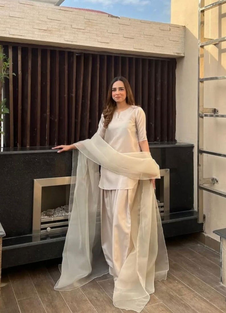 K&ouml;nnte beinhalten: Beigefarbenes zweiteiliges Outfit mit passendem, transparentem Schal. Das Outfit umfasst ein kurz&auml;rmeliges Oberteil und eine locker sitzende Hose. Der Schal ist &uuml;ber den Schultern drapiert. Das Outfit ist aus einem glatten, m&ouml;glicherweise satinartigen Material.