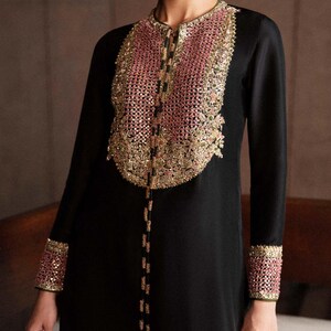 K&ouml;nnte beinhalten: Schwarzes Langarmkleid mit aufwendiger rosa-goldener Stickerei am Ausschnitt und an den Manschetten. Das Kleid hat eine zarte Perlenverzierung auf der Vorderseite, die dem Design einen Hauch von Eleganz verleiht.
