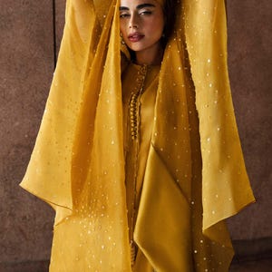 K&ouml;nnte beinhalten: Senfgelbes Ensemble mit einer lang&auml;rmeligen Tunika mit Stehkragen und dekorativen Kn&ouml;pfen. Das Outfit beinhaltet eine passende Kopfbedeckung und einen transparenten, verzierten Schal. Der Schal ist mit kleinen, verstreuten Pailletten verziert.