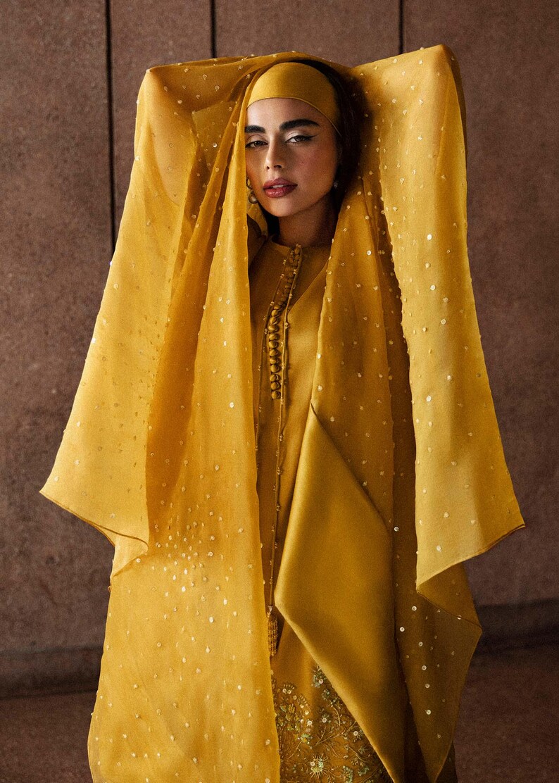 K&ouml;nnte beinhalten: Senfgelbes Ensemble mit einer lang&auml;rmeligen Tunika mit Stehkragen und dekorativen Kn&ouml;pfen. Das Outfit beinhaltet eine passende Kopfbedeckung und einen transparenten, verzierten Schal. Der Schal ist mit kleinen, verstreuten Pailletten verziert.
