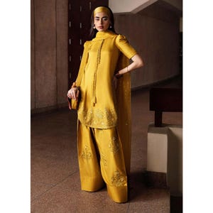 K&ouml;nnte beinhalten: Ein senfgelbes Ensemble mit einer bestickten Tunika, passenden weiten Hosen und einem Kopftuch. Das Outfit umfasst einen transparenten Schal und eine kleine Clutch. Die Kleidung hat einen satinartigen Glanz.
