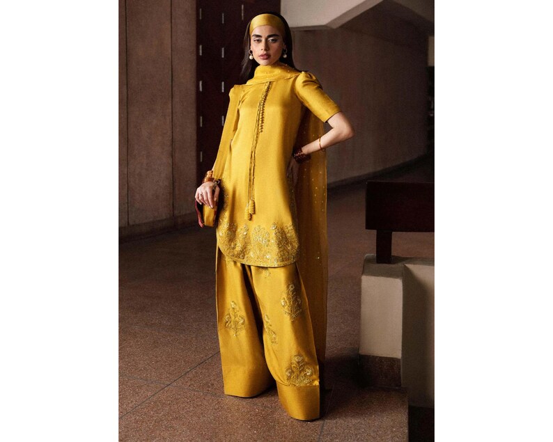 K&ouml;nnte beinhalten: Ein senfgelbes Ensemble mit einer bestickten Tunika, passenden weiten Hosen und einem Kopftuch. Das Outfit umfasst einen transparenten Schal und eine kleine Clutch. Die Kleidung hat einen satinartigen Glanz.