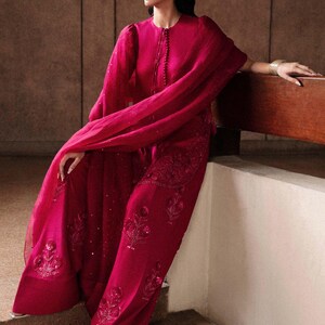 K&ouml;nnte beinhalten: Ein magenta-farbenes Ensemble mit einem lang&auml;rmeligen Oberteil mit rundem Ausschnitt und einer passenden Hose mit weitem Bein. Das Outfit ist mit aufwendiger Stickerei und Pailletten verziert. Ein flie&szlig;ender Schal und ein Stirnband vervollst&auml;ndigen den Look.