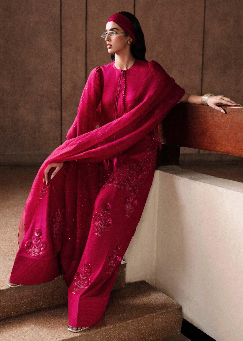 K&ouml;nnte beinhalten: Ein magenta-farbenes Ensemble mit einem lang&auml;rmeligen Oberteil mit rundem Ausschnitt und einer passenden Hose mit weitem Bein. Das Outfit ist mit aufwendiger Stickerei und Pailletten verziert. Ein flie&szlig;ender Schal und ein Stirnband vervollst&auml;ndigen den Look.