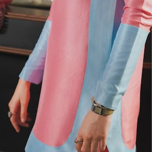 May include: A pink dress with light blue accents. The dress features long sleeves with light blue cuffs and a light blue panel down the front. A silver watch with a green band is visible on the wrist.