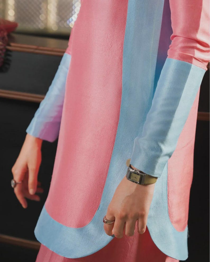 May include: A pink dress with light blue accents. The dress features long sleeves with light blue cuffs and a light blue panel down the front. A silver watch with a green band is visible on the wrist.