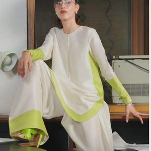 May include: A cream-colored two-piece outfit with lime green accents. The long-sleeved top and wide-leg pants feature green trim. The model is wearing lime green sandals and glasses.