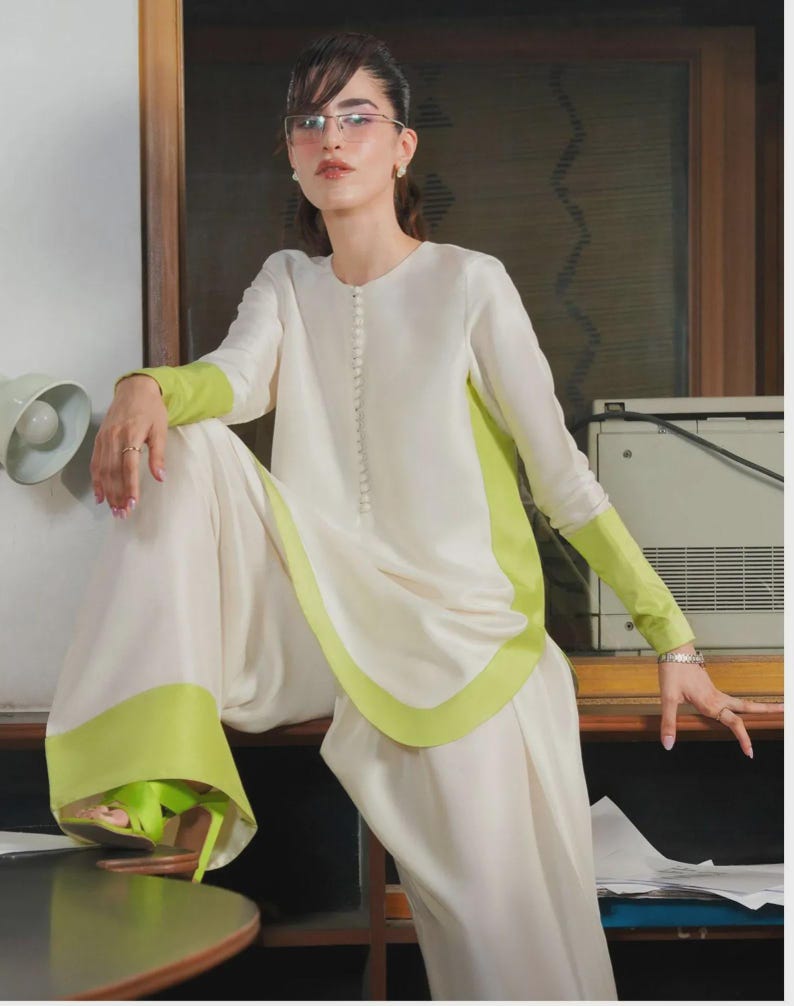 May include: A cream-colored two-piece outfit with lime green accents. The long-sleeved top and wide-leg pants feature green trim. The model is wearing lime green sandals and glasses.