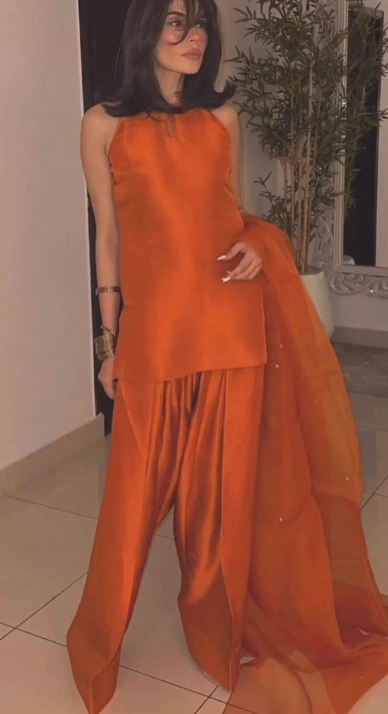 K&ouml;nnte beinhalten: Ein leuchtend orangefarbenes zweiteiliges Outfit. Das Oberteil ist im Neckholder-Stil gehalten, und die Hose ist locker geschnitten. Ein durchsichtiger orangefarbener Schal drapiert das Outfit. Das Ensemble besteht aus gl&auml;nzendem, seidigem Material. Goldene Armb&auml;nder schm&uuml;cken das Handgelenk.