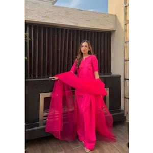 K&ouml;nnte beinhalten: Ein leuchtend pinkfarbenes Salwar-Kameez-Ensemble. Das Outfit umfasst ein Tunika-Oberteil, eine Hose mit weitem Bein und einen transparenten Dupatta-Schal. Das Model tr&auml;gt das Outfit im Freien.