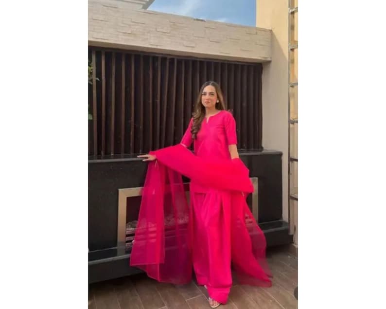 K&ouml;nnte beinhalten: Ein leuchtend pinkfarbenes Salwar-Kameez-Ensemble. Das Outfit umfasst ein Tunika-Oberteil, eine Hose mit weitem Bein und einen transparenten Dupatta-Schal. Das Model tr&auml;gt das Outfit im Freien.