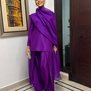 K&ouml;nnte beinhalten: Ein leuchtend violettes Outfit, bestehend aus einem lang&auml;rmeligen Oberteil und einer weiten Hose, wird mit einem passenden Schal kombiniert. Silberner Schmuck und eine kleine Clutch erg&auml;nzen den Look. Das Outfit ist aus einem gl&auml;nzenden Stoff gefertigt.