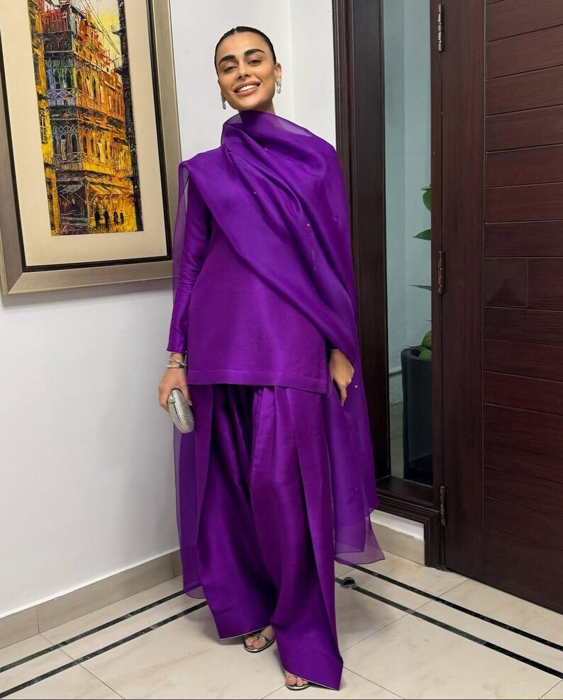 K&ouml;nnte beinhalten: Ein leuchtend violettes Outfit, bestehend aus einem lang&auml;rmeligen Oberteil und einer weiten Hose, wird mit einem passenden Schal kombiniert. Silberner Schmuck und eine kleine Clutch erg&auml;nzen den Look. Das Outfit ist aus einem gl&auml;nzenden Stoff gefertigt.