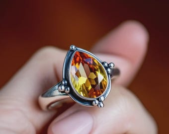 May include: A silver ring featuring a pear-shaped gemstone with yellow, orange, and red hues. The gemstone is faceted and set in a detailed silver band with small decorative accents.