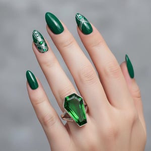 Könnte beinhalten: Eine Hand zeigt einen silbernen Ring mit einem Sarg-förmigen, smaragdgrünen Edelstein. Die Nägel sind in passendem Smaragdgrün lackiert, mit Akzentnägeln mit weißen und silbernen geometrischen Designs. Der Hintergrund ist einfarbig grau.
