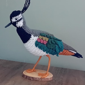 May include: A handcrafted crochet bird sculpture. The bird has a black head with a white and grey face, a grey back, and colourful wings with teal, pink, and green accents. It stands on orange legs attached to a wooden base.