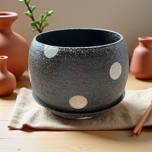 May include: A black ceramic planter with white polka dots. The round pot sits on a matching saucer. The textured surface contrasts with the smooth, circular white accents. The planter is suitable for indoor or outdoor use.