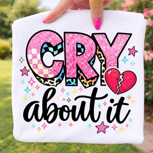 May include: White sweatshirt with the text "CRY about it" in a playful font. The word "CRY" is in pink glitter with checkered and leopard print accents. A broken heart graphic and colorful stars complete the design.