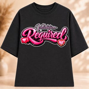 May include: A black short-sleeve t-shirt featuring a graphic print with the text "Validation Required" in a glossy, bubble-letter pink font. The design includes a pink heart-shaped notification icon and a pink heart icon with sparkles.