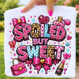 Może przedstawiać: Biała bluza z napisem "SPOILED BUT SWEET" w różowym brokatem, wzorem w cętki i niebieską kratą. Projekt zawiera różowe kokardy, serca, torebkę, szminkę i perfumy.